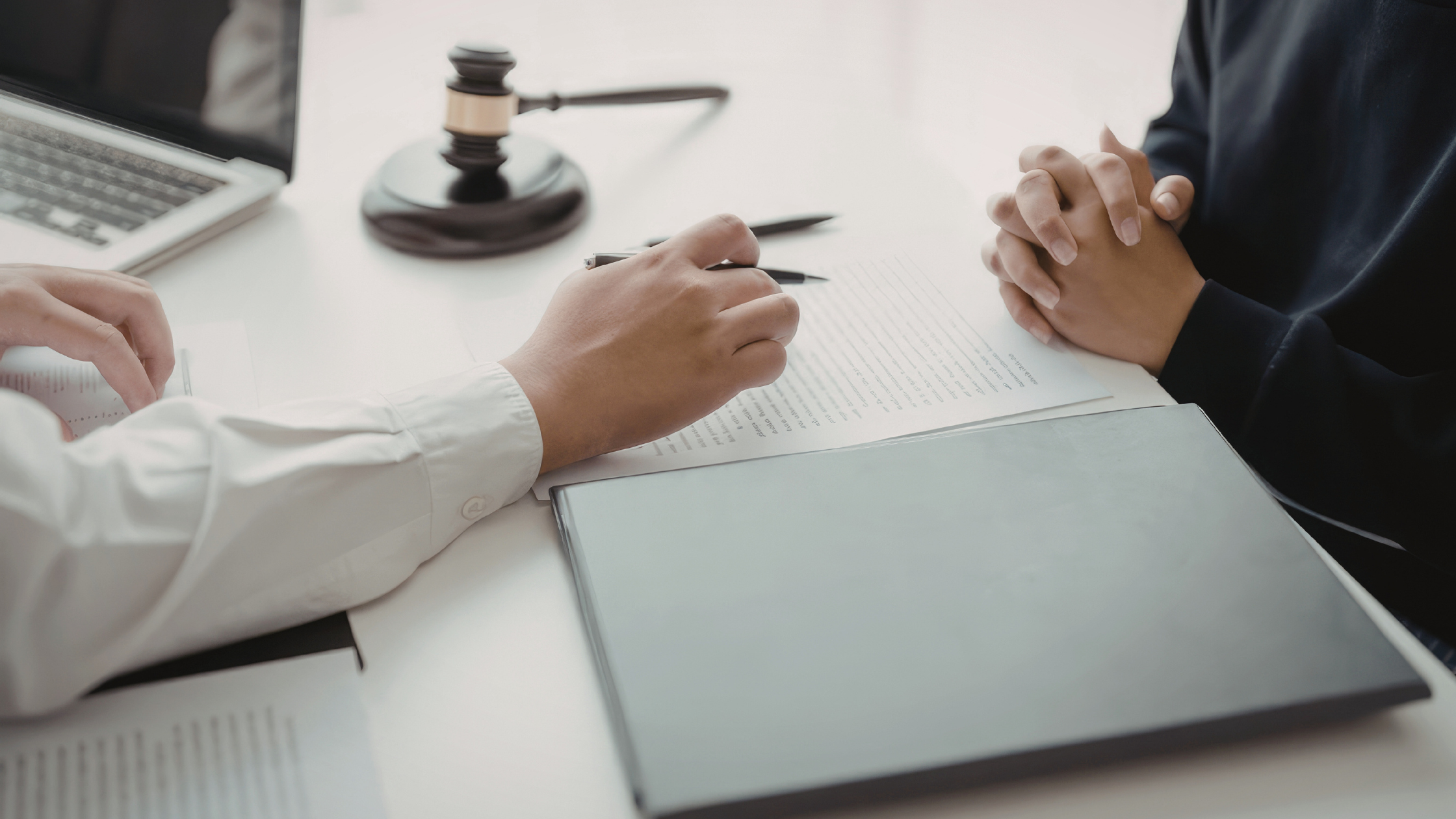 Hand holding pen and pointing toward clasped hands over top of a gray folder with a gavel in the background.